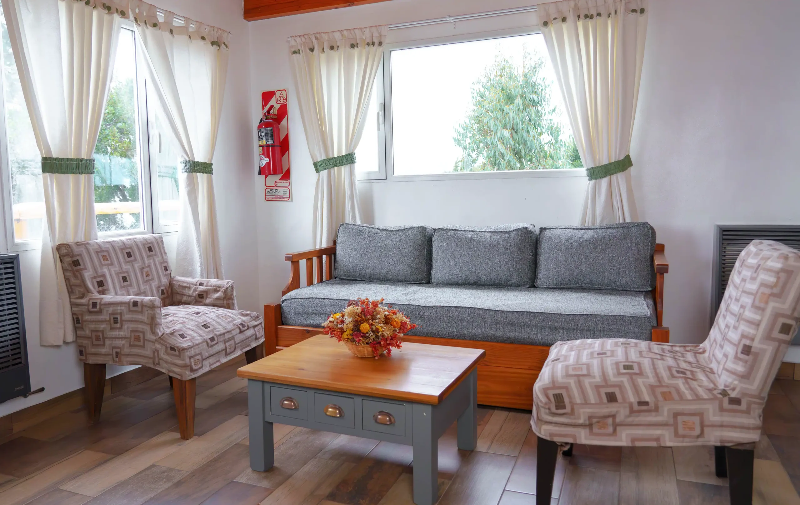 Living con sillón de 3 cuerpos y dos sillones simples a los costados, con mesa ratona, 2 calefactores de tiro balanceado y ventanas, correspondientes a la Casa 2