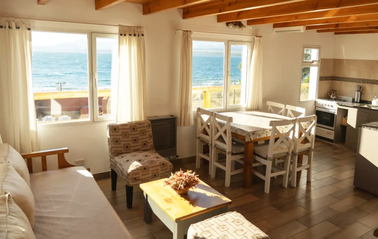 Amplio living comedor con cocina integrada, mesa con 6 sillas, sillón de 3 cuerpos y dos sillones individuales a los costados con mesa ratona y 3 ventanas con vista al lago, correspondientes a la Casa 3