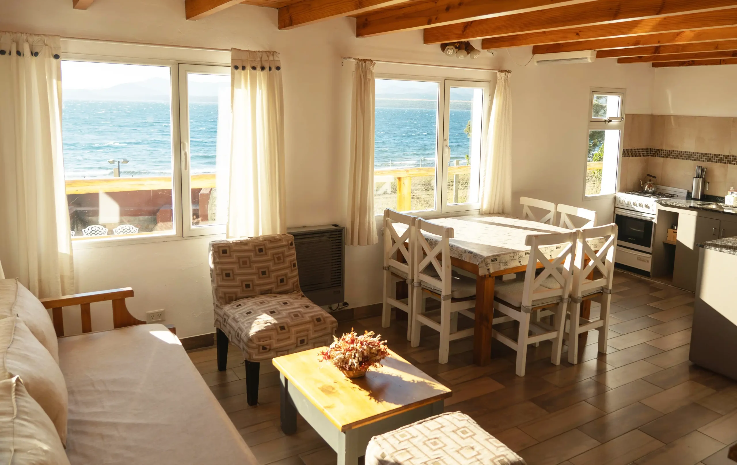 Amplio living comedor con cocina integrada, mesa con 6 sillas, sillón de 3 cuerpos y dos sillones individuales a los costados con mesa ratona y 3 ventanas con vista al lago, correspondientes a la Casa 3