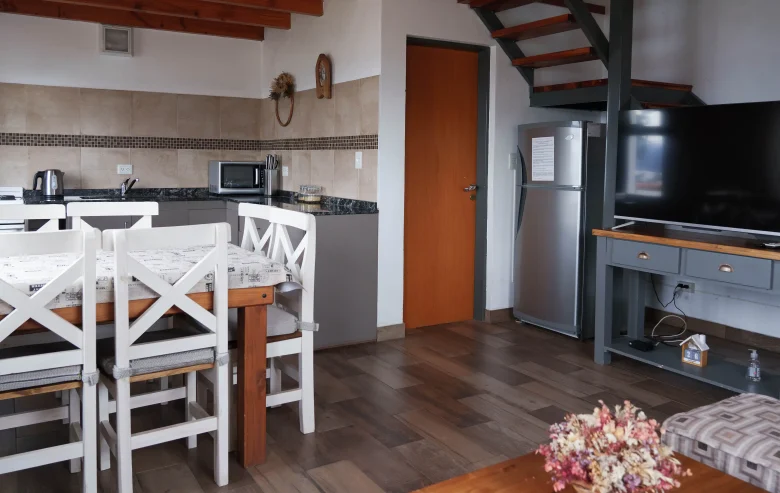 Living comedor con cocina integrada, con microondas y pava eléctrica. Mesa con 5 sillas, heladera grande, mesada con televisor y detrás escaleras para el primer piso, correspondientes a la Casa 3