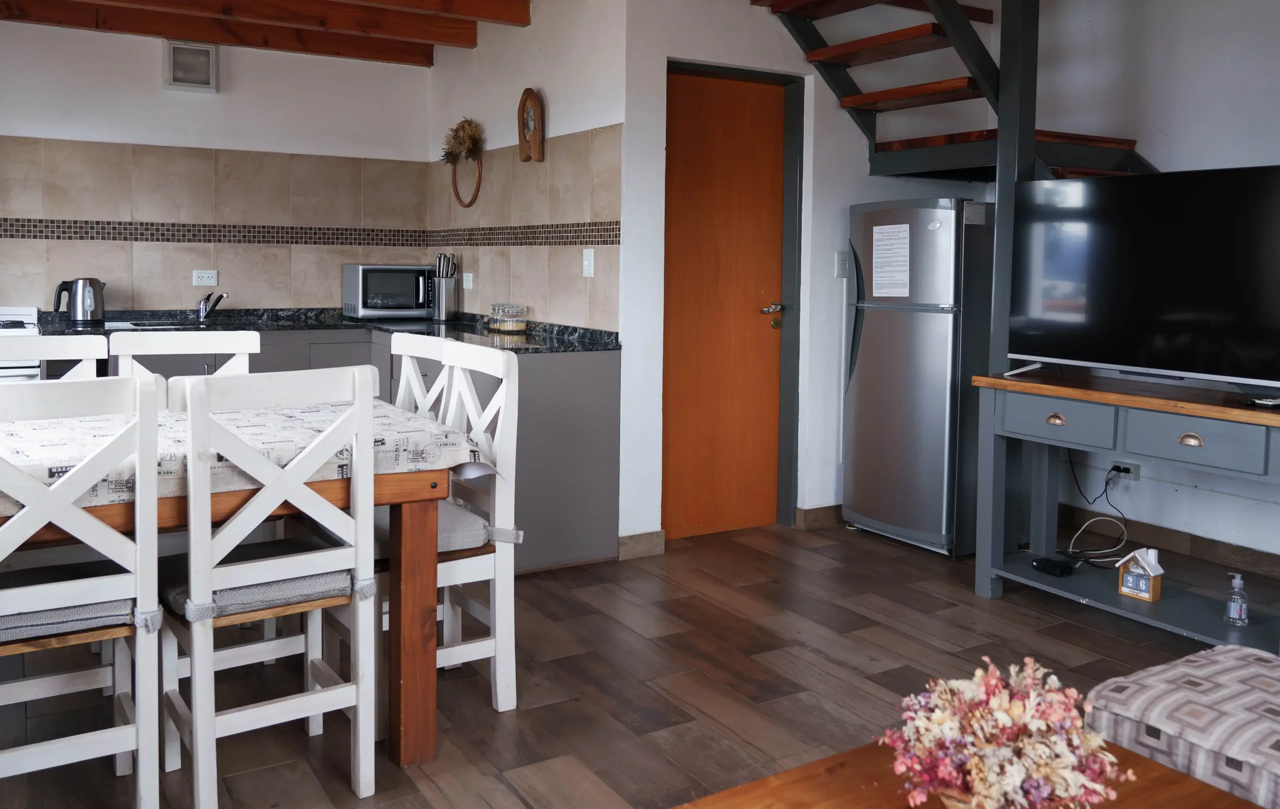 Living comedor con cocina integrada, con microondas y pava eléctrica. Mesa con 5 sillas, heladera grande, mesada con televisor y detrás escaleras para el primer piso, correspondientes a la Casa 3
