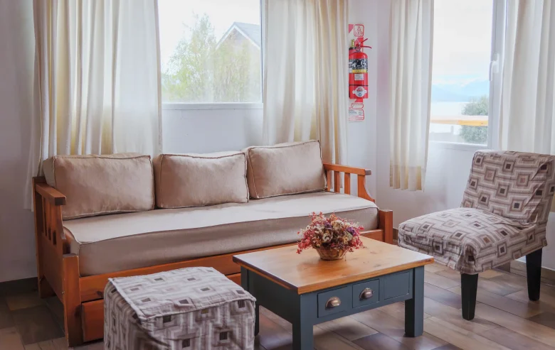 Living con sillón de 3 cuerpos, puff y sillón individual alrededor de mesa ratona y detrás 2 ventanas con vista al lago, correspondiente a la Casa 3