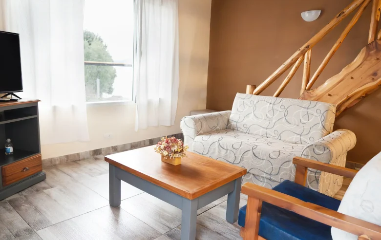 Living con sillón de 2 cuerpos y sillón individual con mesita ratona y mesa con televisor, ventana y cortinas y escalera de madera, correspondiente a la Casa 4