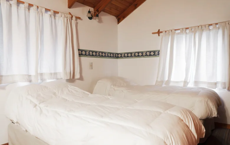 Dormitorio secundario con 2 camas individuales y acolchado de pluma y dos ventanas con cortinas, correspondiente a la Casa 4