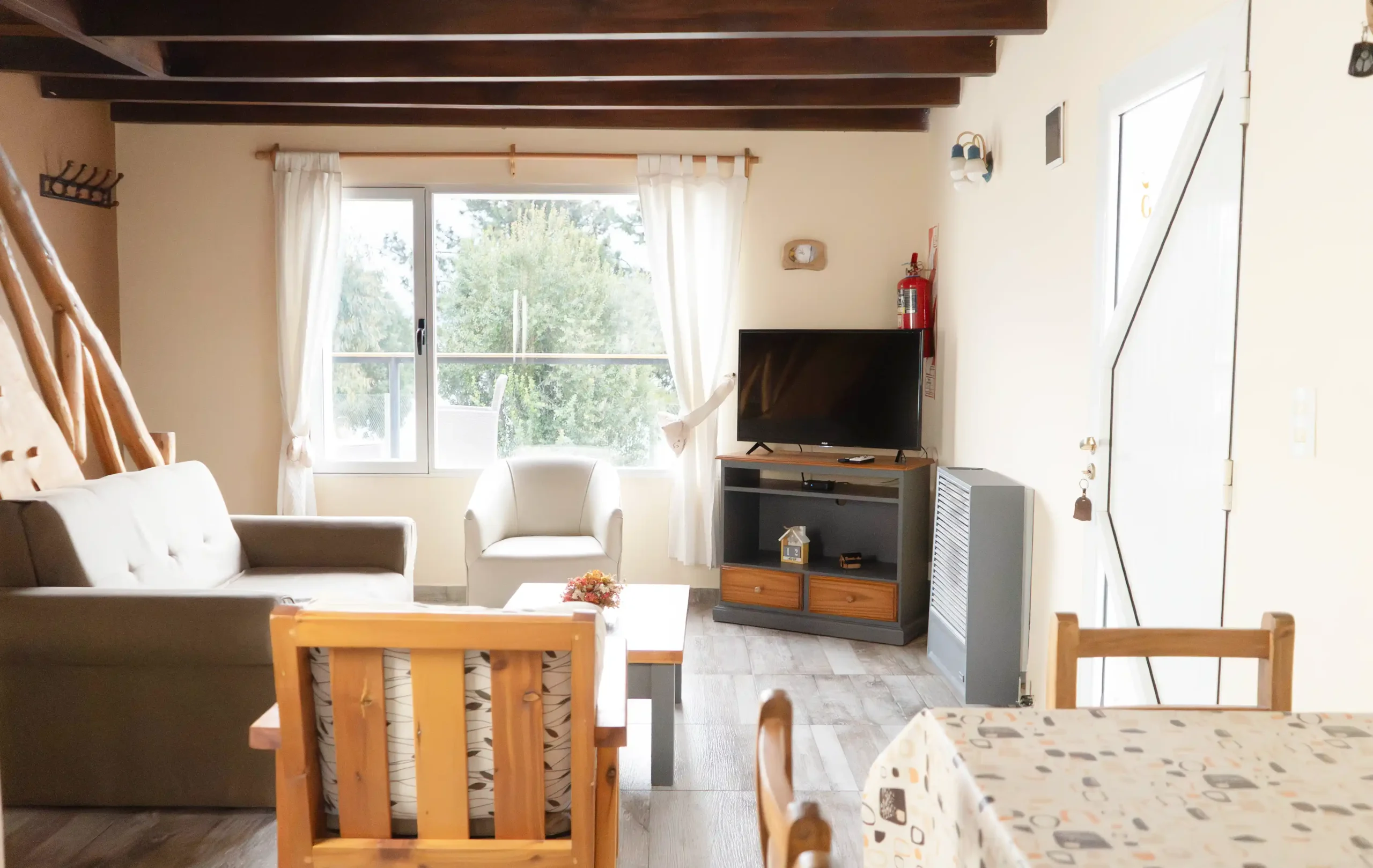 Living comedor, vista parcial de la mesa con 2 sillas, sillón de 3 cuerpos con dos sillones individuales a los costados, mesa ratona, calefactor de tiro balanceado al lado de la puerta, mesa con televisor, ventana con vista al jardín y escalera de madera, correspondiente a la Casa 5